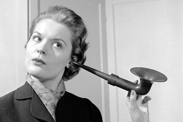 Woman using an ear trumpet to listen at a closed door.