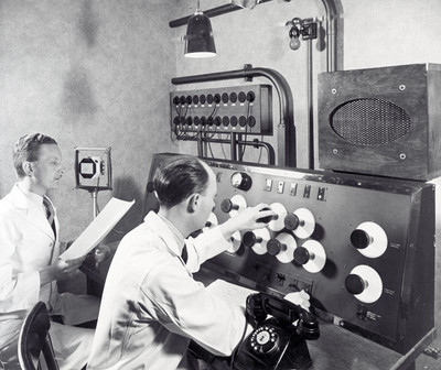 Two men in lab coats working on a large 1950s machine with an array of knobs and dials and a speaker.