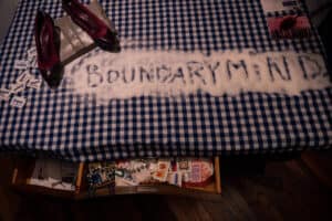 Salt on a table, with 'boundary mind' written in it, plus a pair of shoes, sugar packets and an open drawer containing various ephemera.